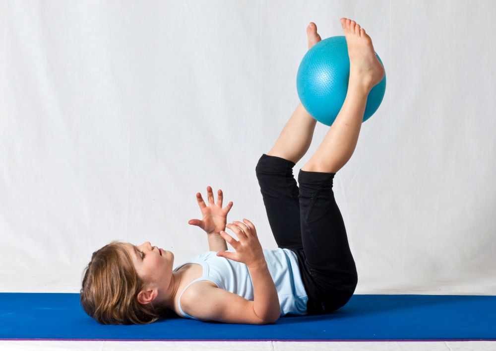 A young girl is lying on her back on a yoga mat holding a gym ball between her legs