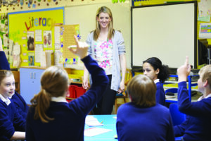 Female teacher at front of class. Pupils raising their hands.